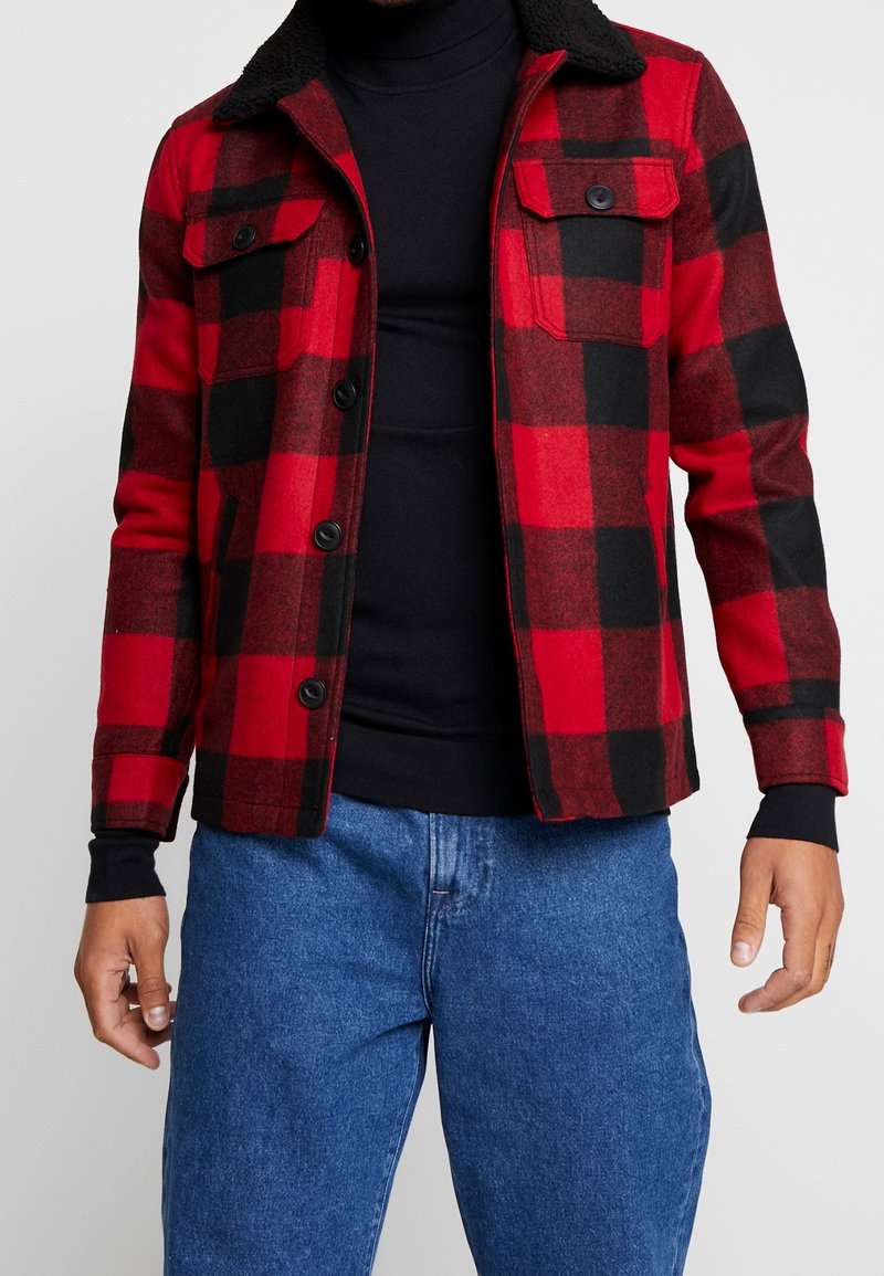 Rød og sort flanneljakke med fleecekrave, frontlukning med knapper og to brystlommer. Bæret over en sort højhalset sweater.