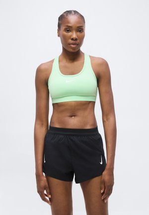 Atletisk kvinde iført en lysegrøn sports-bh og sorte Nike-shorts, der står med neutralt ansigtsudtryk på en ensfarvet lys baggrund.