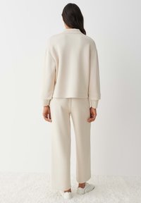 Beige, oversized sweatshirt met een hoge kraag en geribbelde manchetten; gecombineerd met beige broek met wijde pijpen en witte sneakers. Zachte, gladde textuur.