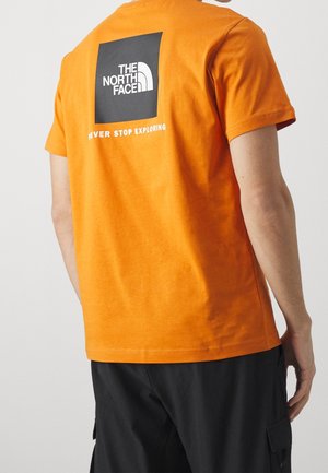 T-shirt in cotone arancione con un logo quadrato nero sul retro, accompagnato da testo bianco: "THE NORTH FACE" e "NEVER STOP EXPLORING". Maniche corte.