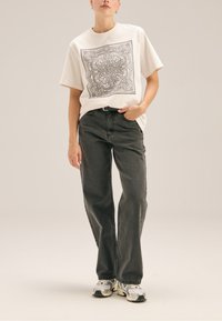 Biała bawełniana koszulka z czarnym wzorem paisley, zestawiona z ciemnoszarymi dżinsami oraz masywnymi szaro-białymi sneakersami.