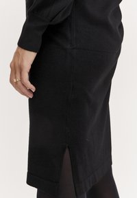 Abito fitted nero in tessuto a maglia con una spaccatura laterale all'orlo, dotato di scollatura e polsini a coste per dettagli di texture e stile.