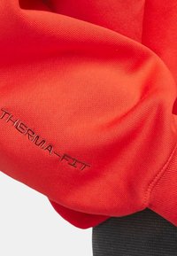 Manga de sudadera Therma-Fit roja con textura acanalada. Presenta un bordado de logo visible en color contrastante. Diseño de tela suave y flexible.