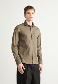 Camisa de manga longa verde-oliva com colarinho abotoados, apresentando um logótipo discreto no peito e botões pretos, em tecido de algodão texturizado.