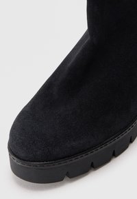 Bottine en suède noir avec une semelle en caoutchouc noir épaisse à bord dentelé et un design de bout arrondi et lisse.