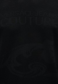Μαύρο υφή ύφασμα με διακριτικό ανάγλυφο κείμενο "VERSACE JEANS COUTURE" και ανάγλυφο σχέδιο που μοιάζει με στιλιζαρισμένο κεφάλι Μέδουσας.