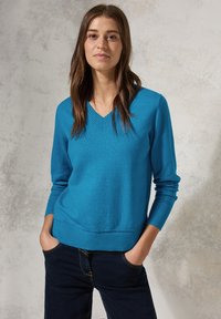Un maglione a v con un tessuto di maglia blu brillante, con polsini e orlo a costine, abbinato a jeans scuri. La superficie è liscia e morbida.