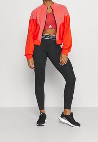 Rød bomberjakke med stribet design, sorte leggings og sorte sportssko. Calvin Klein-logo på taljebåndet, lynlås foran.