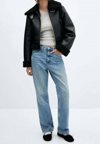 Giacca nera in pelle con collo in montone, top grigio aderente, jeans blu a gamba dritta e ballerine nere. La giacca presenta una cerniera frontale e dettagli con bottoni.