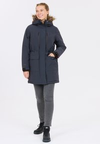 Marineblauer isolierter Parka mit fellgefütterter Kapuze, vorderen Taschen, Kordelzug in der Taille und Reißverschlüssen. Trägt ihn über grauen Jeans und schwarzen Stiefeln.