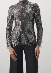 Schwarz-silbernes Langarmoberteil mit Fransen-Detail, hoher Kragen und transparentem Design mit vertikalen Paillettenstreifen.