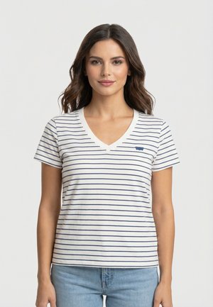 Donna con capelli castani lunghi che indossa una maglietta bianca a V con sottili strisce orizzontali blu scuro e jeans azzurri, in piedi contro uno sfondo semplice.