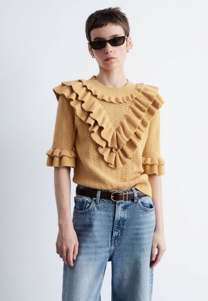 Knit sweater in mustard with large ruffled accents along the shoulders and neckline, paired with light blue high-waisted jeans and a black belt.