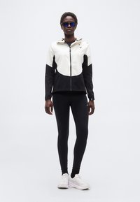 Γυναίκα όρθια φορώντας μαύρο και λευκό μπουφάν με φερμουάρ, μαύρο κολάν, λευκά αθλητικά παπούτσια και γυαλιά ηλίου αθλητικά με μπλε απόχρωση σε λευκό φόντο.