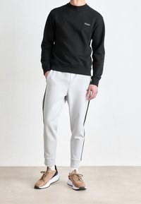 Schwarzer Sweatshirt mit Rundhalsausschnitt, graue Jogginghose mit schwarzen Seitenstreifen und braunen Sportschuhen mit weißen Sohlen. Neutraler Hintergrund.