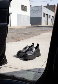 Paire de chaussures à plateforme noires et épaisses sur un trottoir à côté d'une porte de voiture ouverte dans une zone industrielle urbaine.