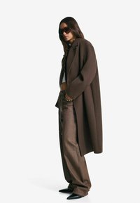 Femme portant un manteau oversized marron, un pantalon large assorti, un crop top blanc, des chaussures noires à bout pointu et de grandes lunettes de soleil sombres, se tenant de profil.