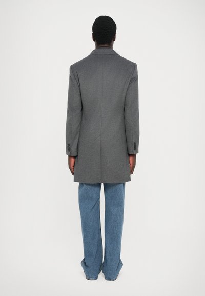 Grå kappa med en strukturerad design, enkel bakventil och stora slag, ihop med vida blå denimbyxor; slät textur.