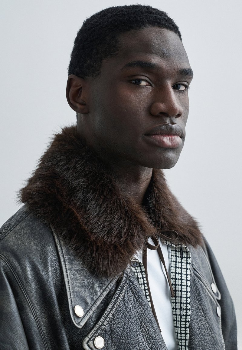 Black leather jacket with a brown fur collar, featuring silver hardware and a checkered inner lining. Close-up of model's profile.