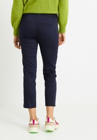 Persona con pantalones azul marino ajustados y cortos, suéter verde y zapatillas blancas con detalles en rosa y verde, de pie frente a un fondo blanco.