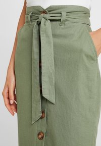 Jupe en lin verte avec une ceinture à nouer à la taille, des boutons sur le devant et deux poches latérales. La texture semble lisse, avec une finition naturelle et décontractée.