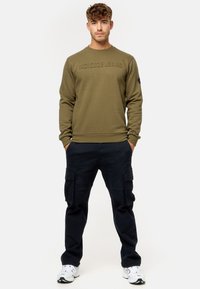 Sudadera verde oliva con el logo texturizado "INDICODE JEANS" y cuello redondo. Combinada con pantalones cargo azul marino que cuentan con múltiples bolsillos y puños.