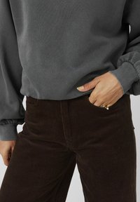 Sweat-shirt gris avec des manches froncées, associé à un pantalon en velours côtelé marron avec des côtes verticales. Main reposant dans la poche avec un détail de bague visible.