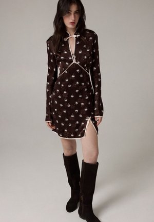 Un mannequin pose devant un fond de studio uni, vêtu d’une mini-robe sombre à fleurs bordée d’un passepoil crème et de bottes noires montant jusqu’aux genoux.