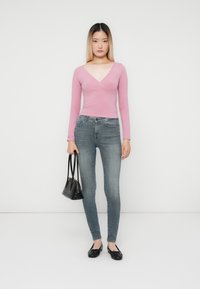 Fiatal nő áll, előre nézve, rózsaszín hosszú ujjú felsőt, elhasználódott skinny farmert, fekete balettcipőt visel, és egy kis fekete táska van a kezében.
