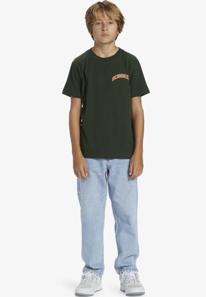 Jongen met lichtbruin haar staat in een donkergroene T-shirt met korte mouwen, lichtblauwe jeans en witte sneakers tegen een witte achtergrond.