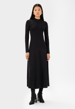 Zwart langwerpig-jurk met lange mouwen, geribbelde textuur, aansluitend lijfje, uitlopende rok, boven de enkel lengte. Model draagt hielen soklaarzen.