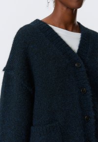 Dunkelblaues Strickcardigan mit V-Ausschnitt, großen Knöpfen und frontalen Taschen. Der Stoff hat ein weiches, strukturiertes Aussehen und eine lockere Passform.