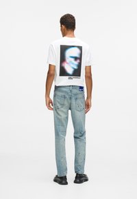 Λευκό μπλουζάκι με πολύχρωμη εκτύπωση στο πίσω μέρος, συνδυασμένο με ανοιχτόχρωμο μπλε τζιν που έχει λεκέδες από μπογιά και μαύρα παπούτσια.