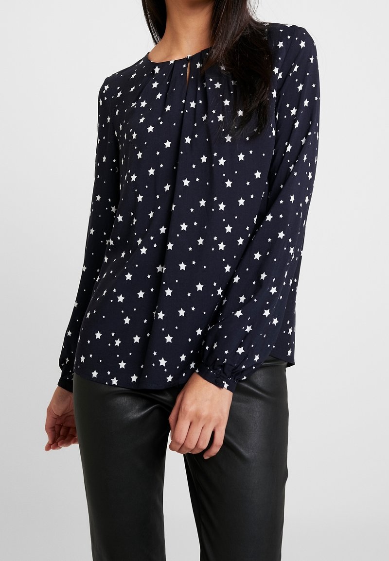 Femme portant un chemisier bleu marine à manches longues avec des motifs d'étoiles blanches et un pantalon en cuir noir sur un fond uni clair.