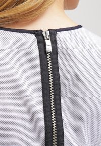 Bílo-černá látka s viditelným kovovým zipem na zadní straně, ohraničená texturovaným černým lemem.