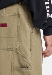 Beige kanvas cargo bukser med en stor lomme og rød merkevaretag. Har synlige sømmer, en trykknapp på lommen og en rett passform.