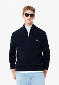 Navy blue zip-neck sweater in soft knit material with ribbed cuffs and hem, accented by a small green logo on the left chest. Worn over a white turtleneck.