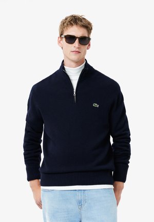 Navy blue zip-neck sweater in soft knit material with ribbed cuffs and hem, accented by a small green logo on the left chest. Worn over a white turtleneck.
