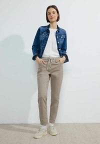 Chaqueta de denim, blusa blanca ajustada, pantalones beiges claros con cordón y zapatillas beige. La modelo posa con las manos en los bolsillos contra un fondo neutro.