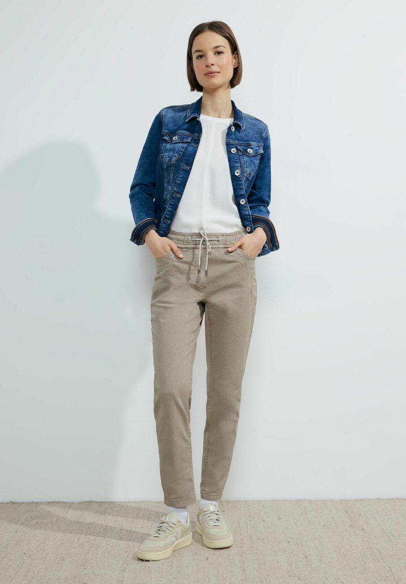 Chaqueta de denim, blusa blanca ajustada, pantalones beiges claros con cordón y zapatillas beige. La modelo posa con las manos en los bolsillos contra un fondo neutro.