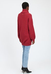 Rød oversized sweater med høj krave, prydet med et tekstureret strikkemønster. Bæres med lyseblå jeans og sorte hælede støvler.