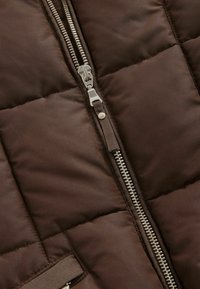 Veste rembourrée marron avec une fermeture éclair diagonale, quincaillerie en argent brillant, texture matelassée et poches latérales avec une finition lisse.