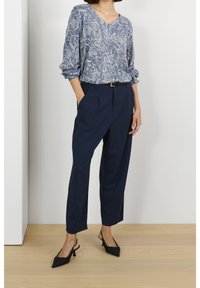 Blouse bleue à motifs avec un col en V et des manches retroussées, associée à un pantalon taille haute bleu marine. Des escarpins noirs à bout pointu complètent la tenue.