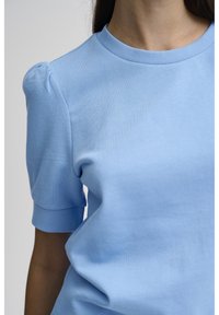 Kvinna iklädd en ljusblå kortärmad tröja med puffärmar och ribbad rund halsringning, avkortad vid halsen och mitt på bålen.