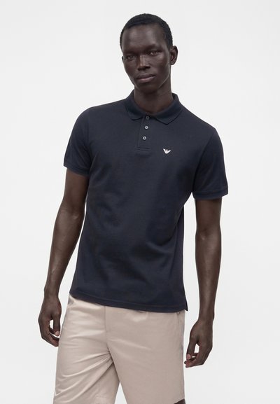 Homme portant un polo noir à manches courtes et un short beige, debout devant un fond blanc uni.