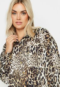 Koszula w zoombie w kolorze panterki z miękkiego materiału, z długimi rękawami i kieszenią z przodu. Złote guziki kontrastują z ciemnym tłem.