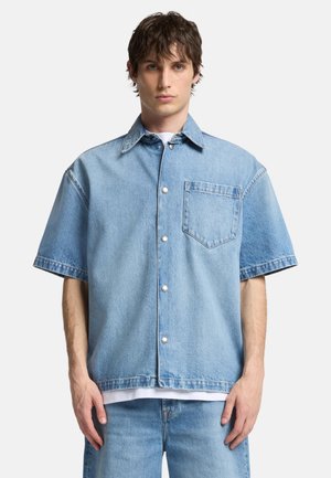 Cămașă din denim de culoare albastră deschisă, cu mâneci scurte, guler ascuțit, buzunar frontal și nasturi cu capsenă argintie. Prezintă o croială lejeră și o textură moale.