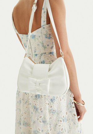Mujer con vestido floral claro que lleva un bolso blanco de hombro con un gran detalle de lazo y cremallera dorada a su lado.