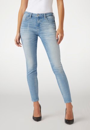 Vrouw draagt lichtblauwe skinny jeans en zwarte hoge hakken, staand tegen een effen witte achtergrond.