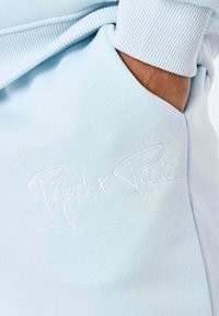 Lichtblauw sweatshirtstof met een zakje waarop de tekst "Project x Paris" is geborduurd. Zachte textuur met geribbelde randafwerking.